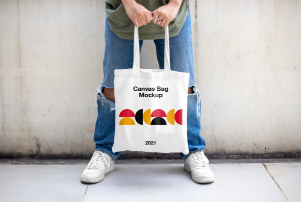Men in bright jeans standing against the wall and holding a Canvas Bag Mockup in both hands.