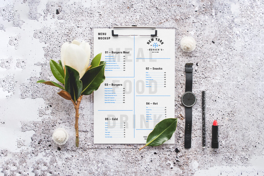 Seen above, a Menu Mockup lies on the concrete with accessories like watch, lipstick and flower.