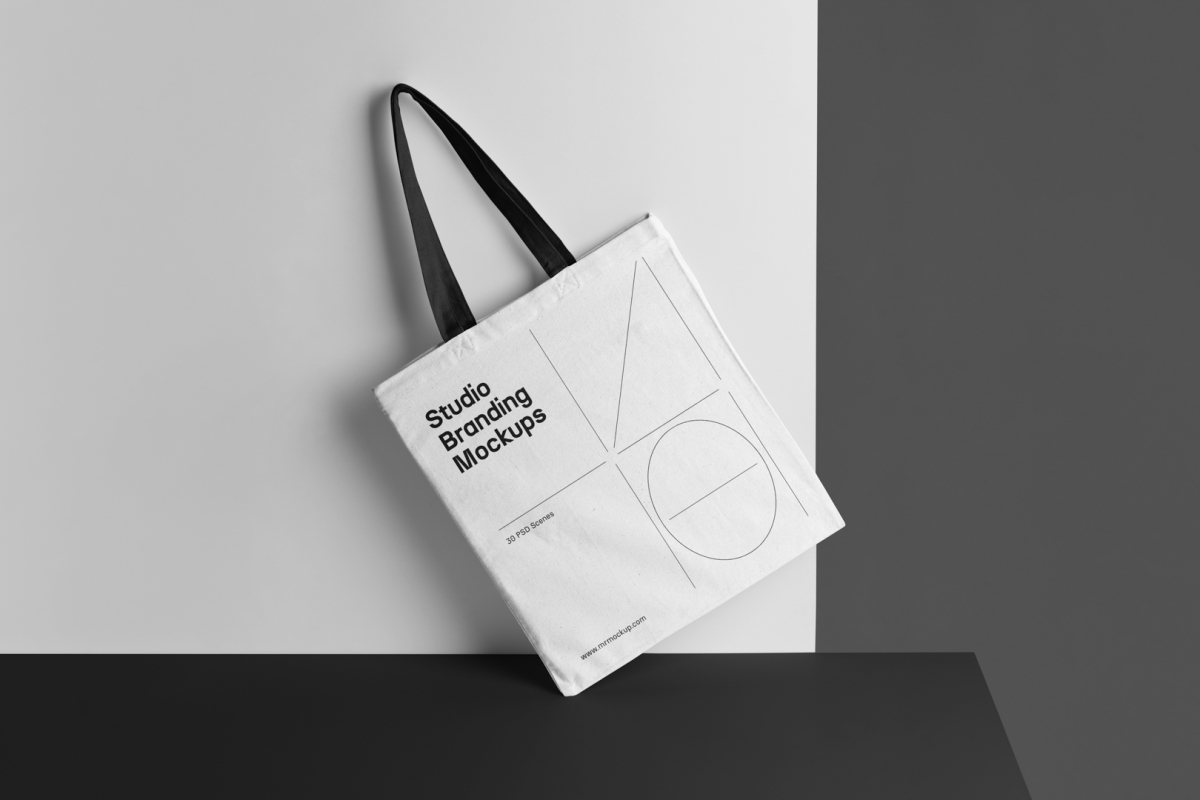 White Canvas Bag Mockup with black handle standing on the black table and is seen from the front.