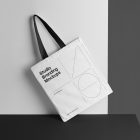 White Canvas Bag Mockup with black handle standing on the black table and is seen from the front.