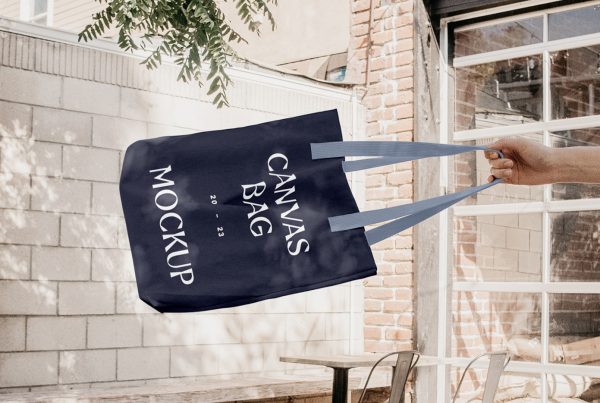 Front view, Free Shopping Tote Bag Mockup with navy design colors and white typography in man's hand against the rest of the restaurant yard in the background.