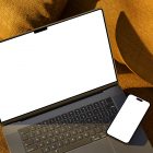Seen from above, space gray blank MacBook Pro Mockup and black color blank iPhone 14 Pro Mockup laying on the reddish yellow couch.