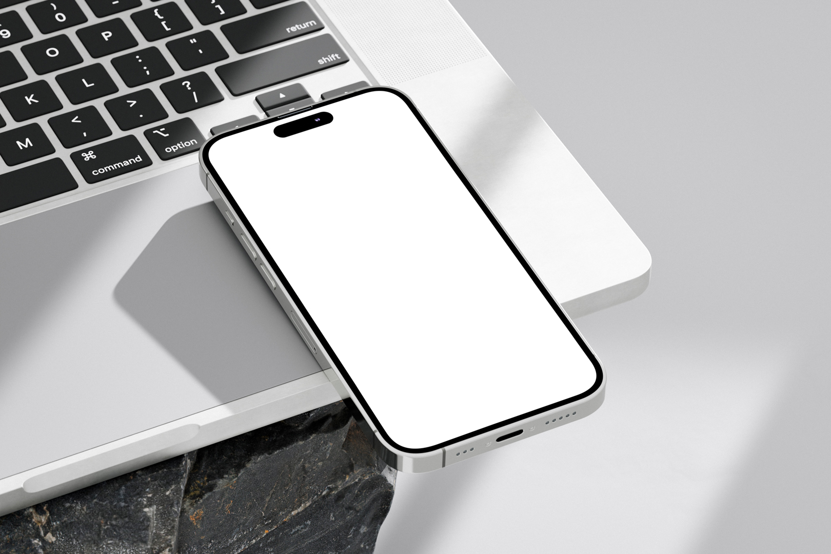 Seen from above, blank iPhone 14 Pro Mockup laying on the MacBook standing on the big rock against a bright gray background.