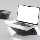 Front view, blank MacBook Pro Mockup standing on the big piece of rock laying on the bright gray floor against second gray/black rock and gray wall in the background.