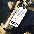 Seen from above, iPhone 14 Pro Mockup with white design laying on the sunny rocks on the beach against water in the background.