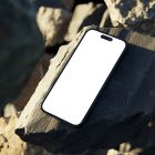 Seen from above, blank iPhone 14 Pro Mockup laying on the sunny rocks on the beach against water in the background.