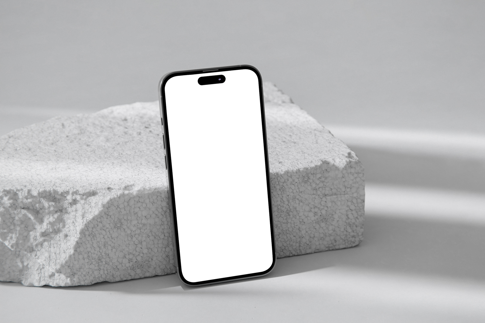 Front view, blank iPhone 14 Pro Mockup based on the piece of a styrofoam and standing on the white floor against white wall.