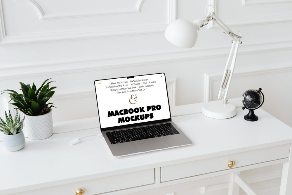 Front view MacBook Pro Mockups with white design laying on the white wooden desk with gold elements, with a white desk lamp, flowers, and black mini globe. Everything is against a white wall.