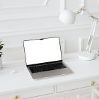 Front view, blank MacBook Pro Device Mockups laying on the white wooden desk with gold elements, with a white desk lamp, flowers, and black mini globe. Everything is against a white wall.