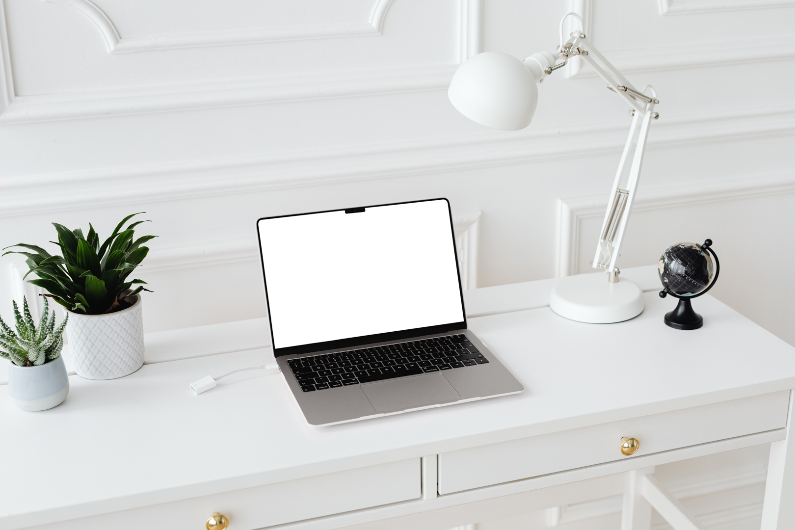 Front view, blank MacBook Pro Device Mockups laying on the white wooden desk with gold elements, with a white desk lamp, flowers, and black mini globe. Everything is against a white wall.