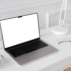 Front view, blank MacBook Pro Device Mockups laying on the white wooden desk with gold elements, with a white desk lamp and white cup, everything is against a white wall.