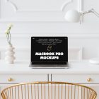 Front view MacBook Pro Mockups with black design laying on the white wooden desk with gold elements, with a white desk lamp and modern vase. Before the white desk standing gold desk chair. Everything is against a white wall.