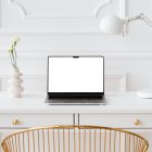 Front view, blank MacBook Pro Device Mockups laying on the white wooden desk with gold elements, with a white desk lamp and modern vase. Before the white desk standing gold desk chair. Everything is against a white wall.