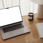 Front view, blank MacBook Pro Device Mockups laying on the wooden desk with a candle, magazine, and modern desk lamp against a white curtain.