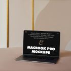 Front view MacBook Pro Mockups with black design laying on the beige table against the wall with white, gold and beige decoration in the background.