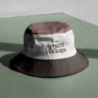 Front view, Bucket Hat Mockup in three colors. Dark brown peak, white, and burgundy laying on the green concrete against white wall.