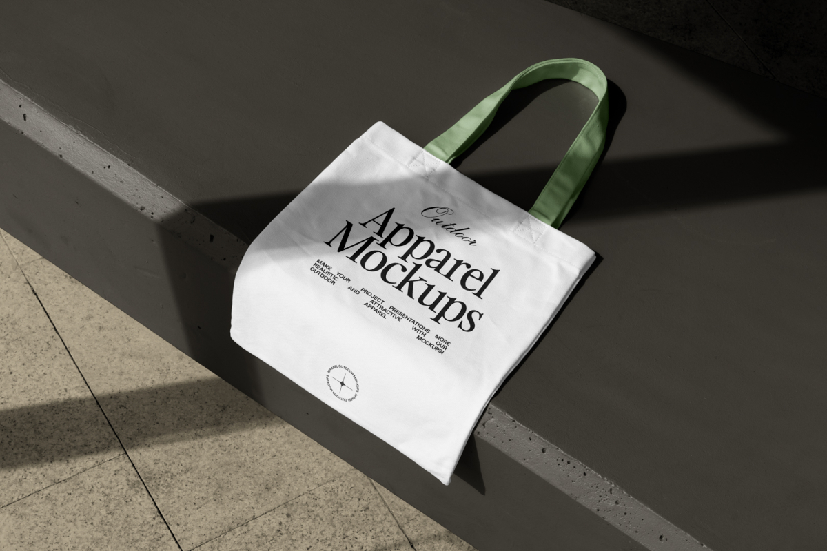 Seen from above, White Canvas Bag Mockup with green handle laying on the dark beige concrete against the marble floor.