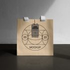 Front view, creamy color Paper Bag Mockup with white handle and craft figure label standing on the dark beige concrete against the white wall.