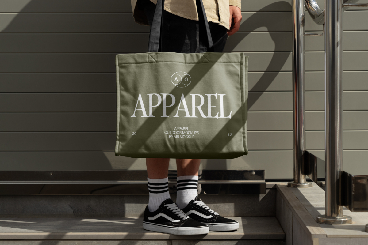 Front view, Men in vans shoes wearing Green Big Canvas Bag Mockup with a black handle and white typographic design on it, against a rotten green wall.