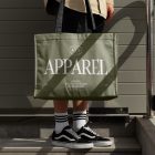 Front view, Men in vans shoes wearing Green Big Canvas Bag Mockup with a black handle and white typographic design on it, against a rotten green wall.