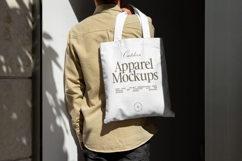 Front view, guy with his back turned in creamy shirt wearing a white Canvas Bag Mockup with a gold typographic design on it against building.