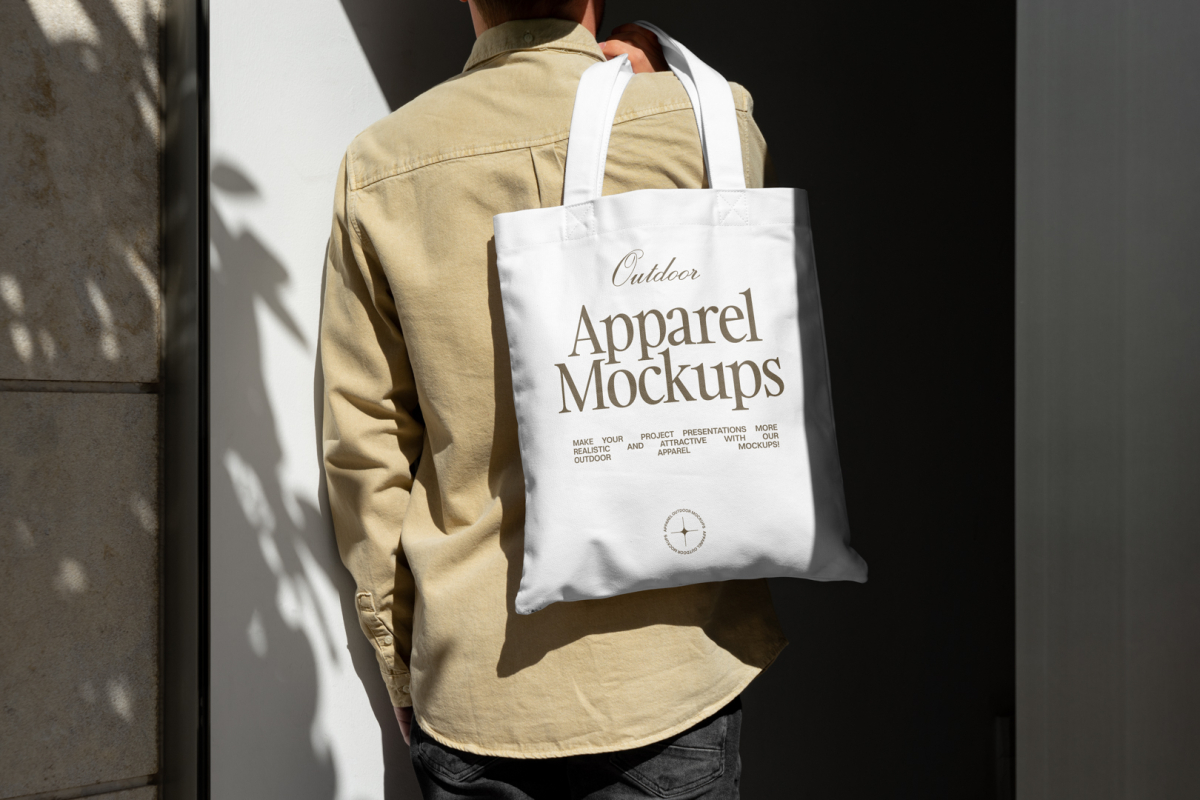 Front view, guy with his back turned in creamy shirt wearing a white Canvas Bag Mockup with a gold typographic design on it against building.