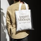 Front view, guy with his back turned in creamy shirt wearing a white Canvas Bag Mockup with a gold typographic design on it against building.