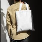 Front view, guy with his back turned in creamy shirt wearing a White Canvas Bag Mockup against building.