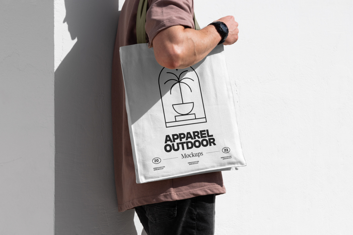 Front view, someone standing sideways and leaning against a white wall in a burgundy t-shirt wearing a white Canvas Bag Mockup with a green handle and black typographic design on it.