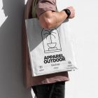 Front view, someone standing sideways and leaning against a white wall in a burgundy t-shirt wearing a white Canvas Bag Mockup with a green handle and black typographic design on it.