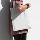 Front view, someone standing sideways and leaning against a white wall in a burgundy t-shirt wearing a White Canvas Bag Mockup.