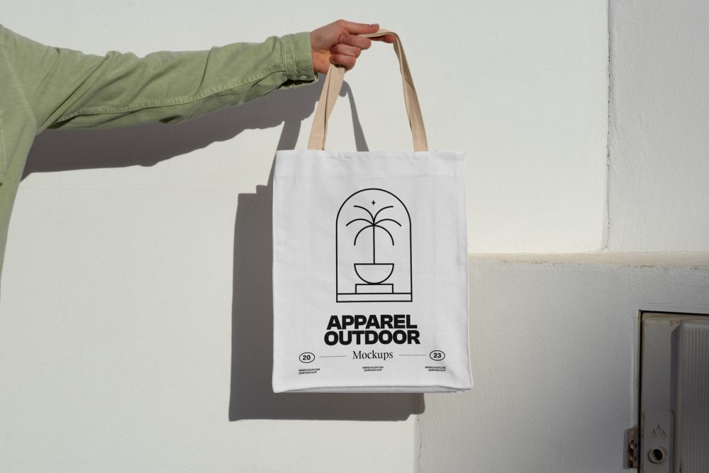 Front view, arm holding a white Canvas Bag Mockup with creamy handle and black typographic design on it against on the white wall.