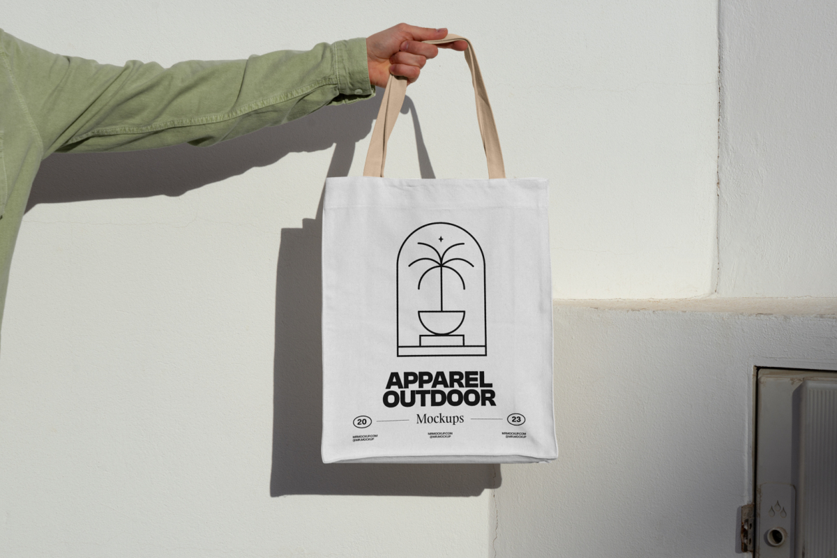 Front view, arm holding a white Canvas Bag Mockup with creamy handle and black typographic design on it against on the white wall.