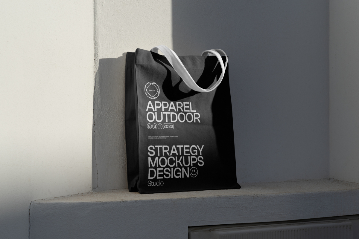 Front view, Black Canvas Bag Mockup with white handle and white typographic design standing on the white windowsill against on the white wall in shadow.