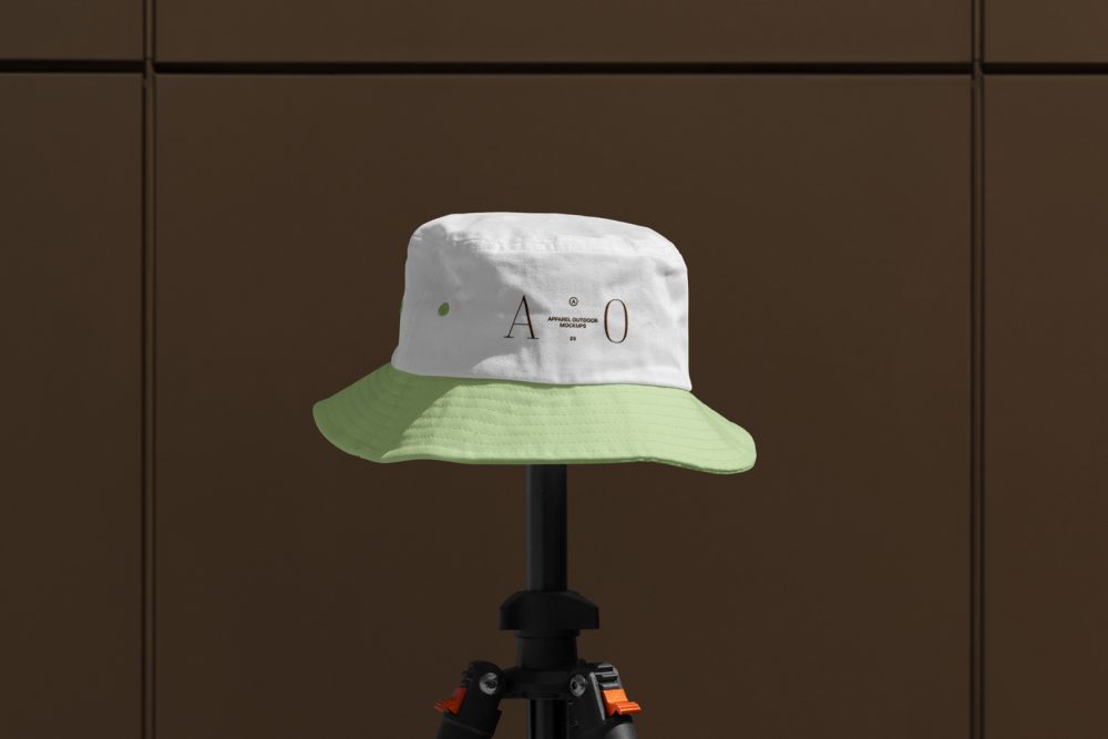 Front view, Bucket Hat Mockup with green peak and white in the upper part of the cap with embroidery of a gold typographic design effect is mounted on a black tripod against on the dark brown wall.