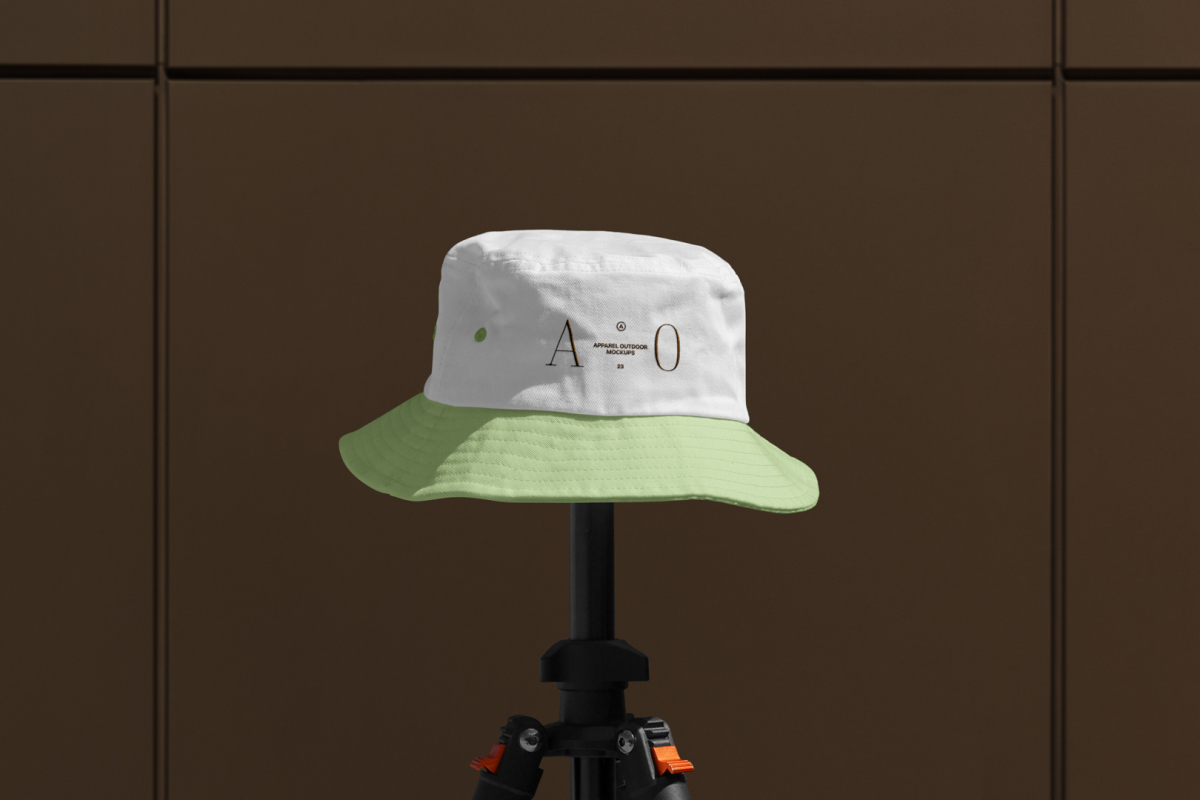 Front view, Bucket Hat Mockup with green peak and white in the upper part of the cap with embroidery of a gold typographic design effect is mounted on a black tripod against on the dark brown wall.