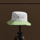 Front view, Bucket Hat Mockup with green peak and white in the upper part of the cap with embroidery of a gold typographic design effect is mounted on a black tripod against on the dark brown wall.