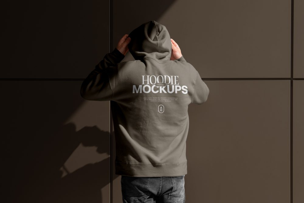 Front view, a man with his back turned, wearing a white gray Hoodie Mockup standing against on the dark gray wall.