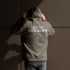 Front view, a man with his back turned, wearing a white gray Hoodie Mockup standing against on the dark gray wall.