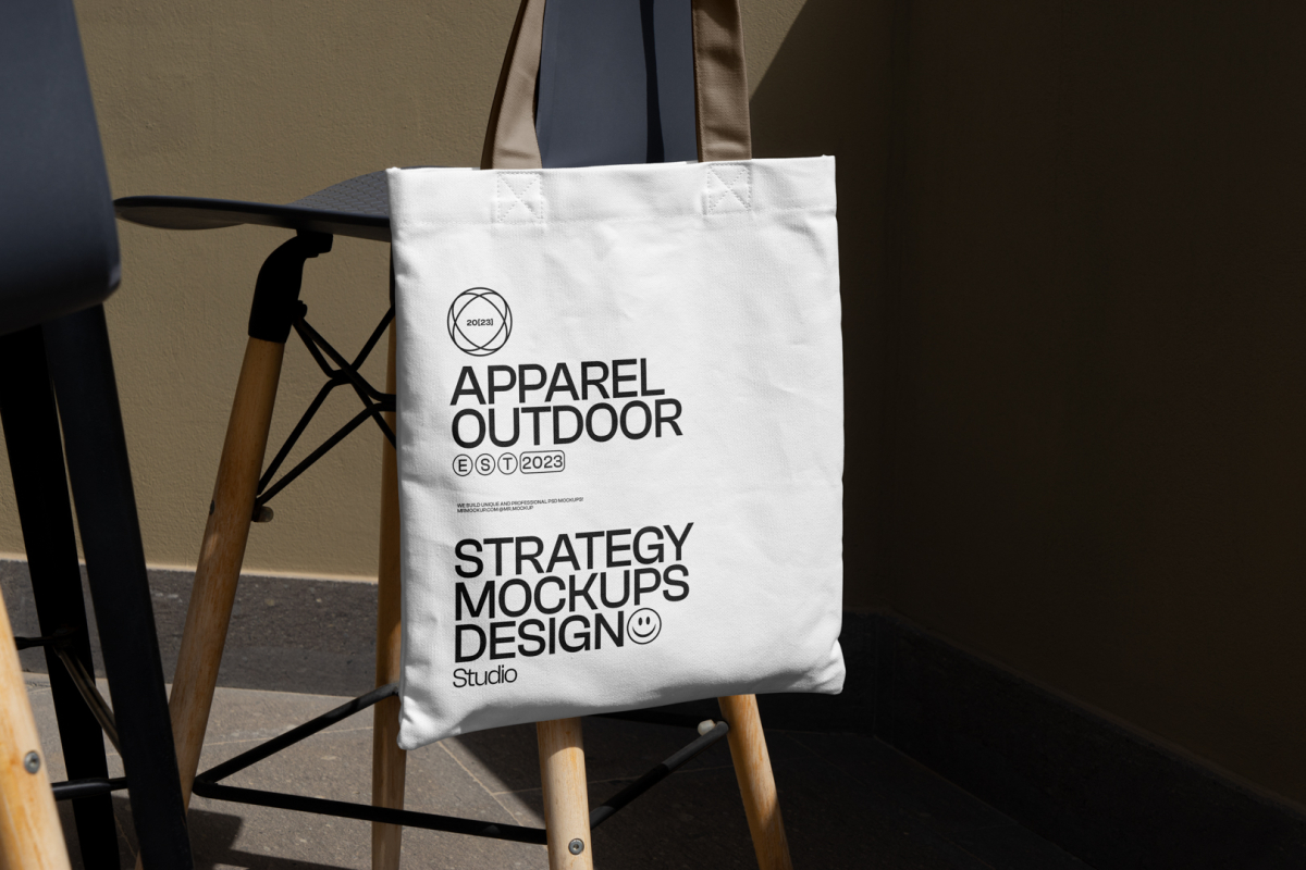 Front view, White Canvas Bag Mockup with beige handle and black typographic design hanging on the chair against gold wall.