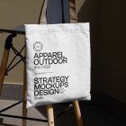 Front view, White Canvas Bag Mockup with beige handle and black typographic design hanging on the chair against gold wall.