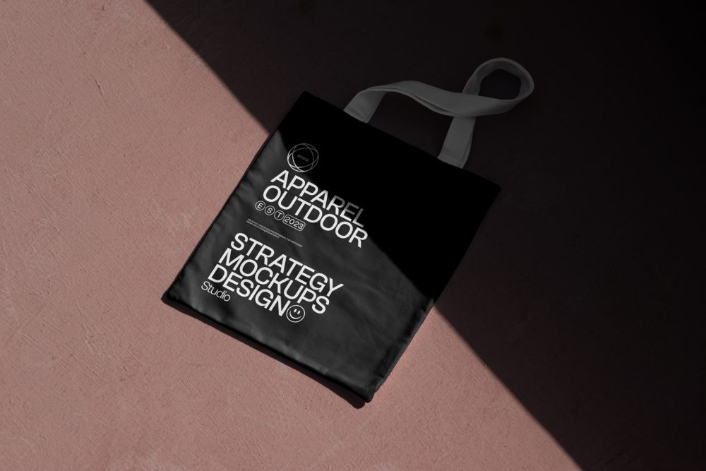 Seen from above, a black Canvas Bag Mockup with a white handle and white typographic design on it laying on the burgundy floor.