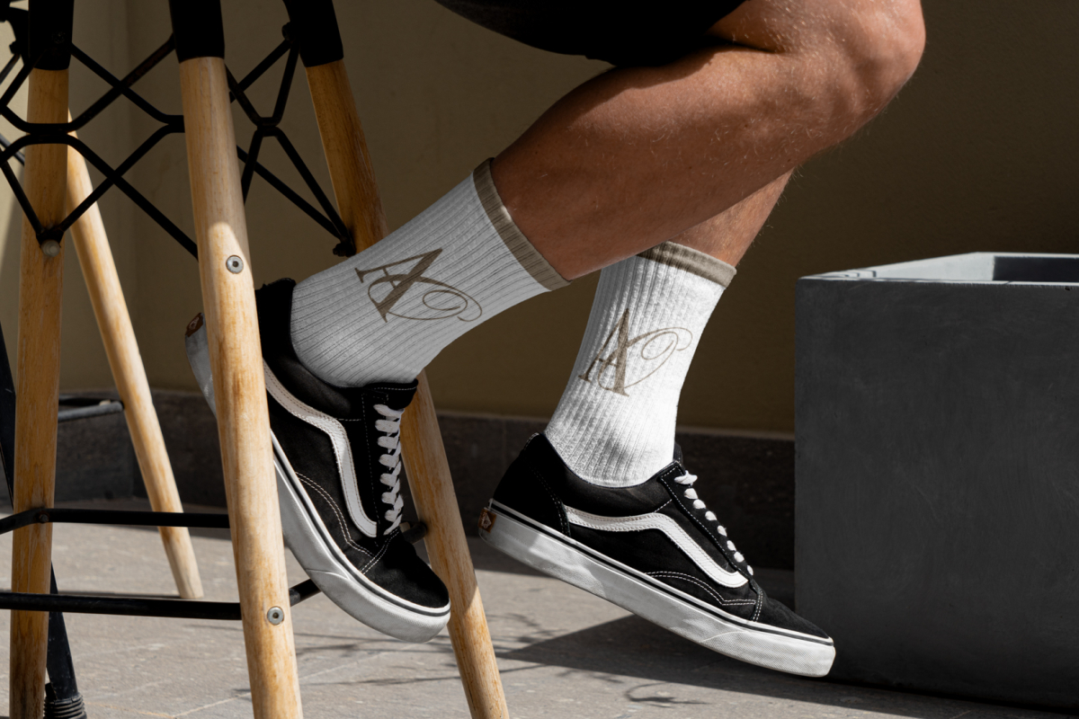 Front view, Men wearing vans shoes and white Socks Mockup with gold typographic design on it sitting on a chair against a gold wall.