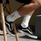 Front view, Men wearing vans shoes and white Socks Mockup with gold typographic design on it sitting on a chair against a gold wall.