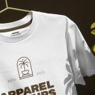 Front view, White T-Shirt Mockup hanging on a lime hanger and gold typographic design on it against a dark brown wall and monster flower.