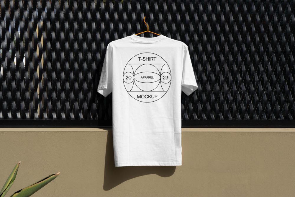 Front view, white T-Shirt Mockup with a black typographic logo on it hanging on an orange hanger attached to a black fence.
