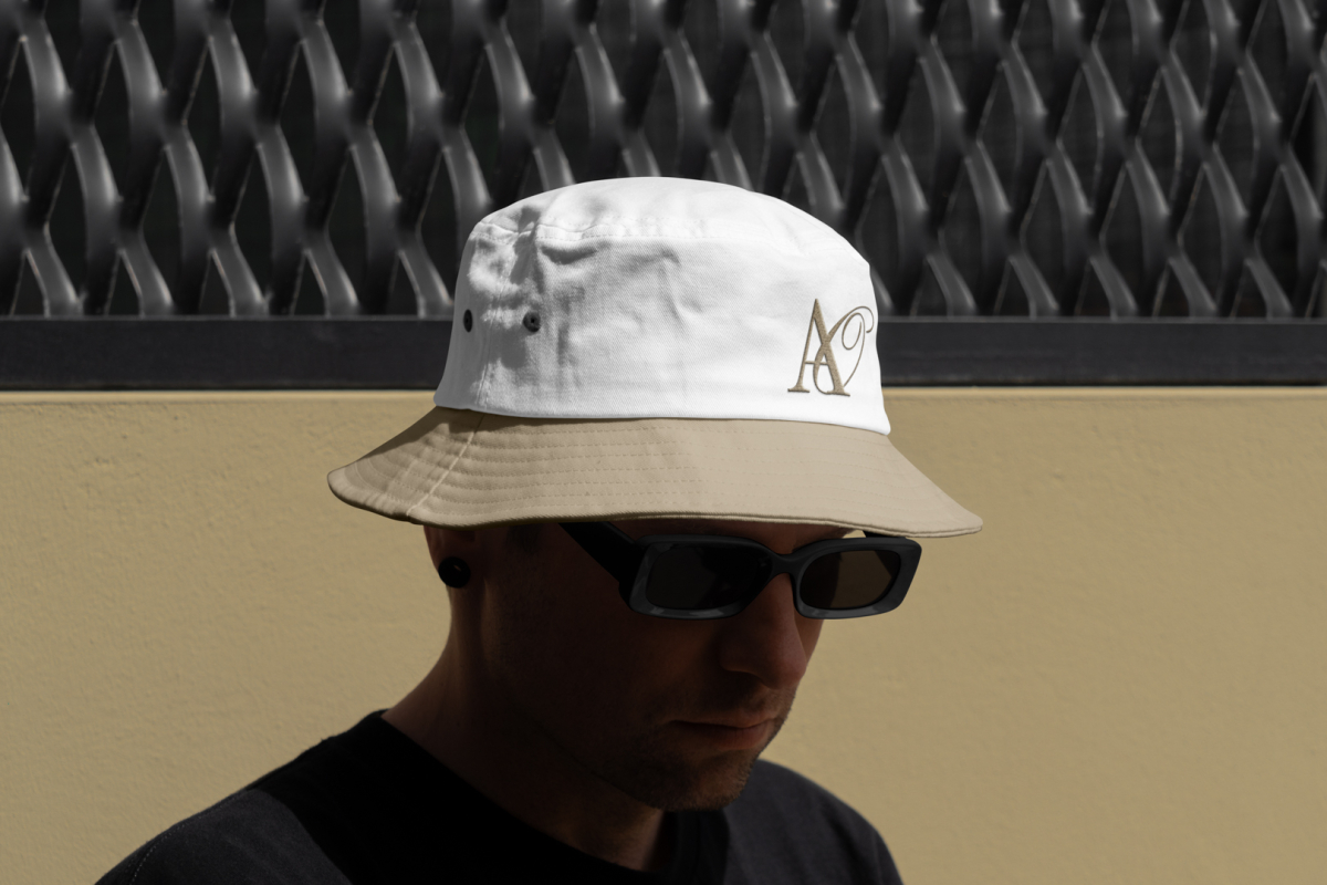 Front view, Men wearing beige, white Bucket Hat Mockup with embroidery of a gold typographic design against a black fence.