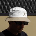 Front view, Men wearing beige, white Bucket Hat Mockup with embroidery of a gold typographic design against a black fence.