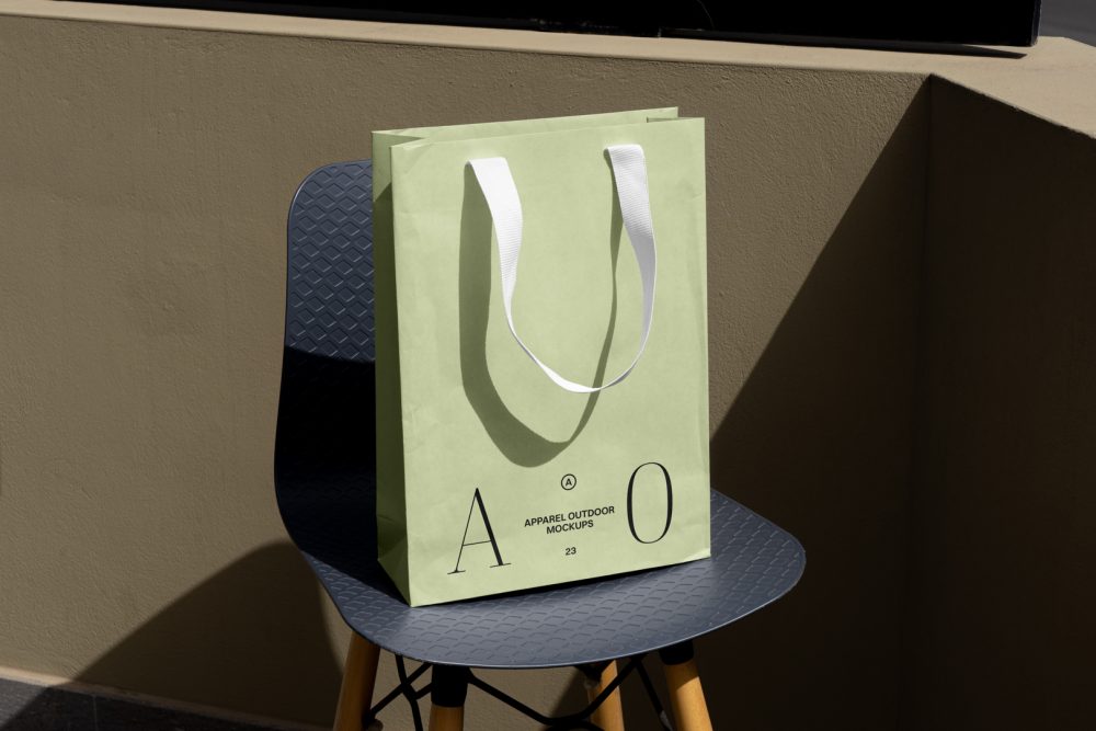 Front view, luxury green Paper Bag Mockup with a white handle and black typographic design on it standing on the navy chair with wooden legs against beige wall.