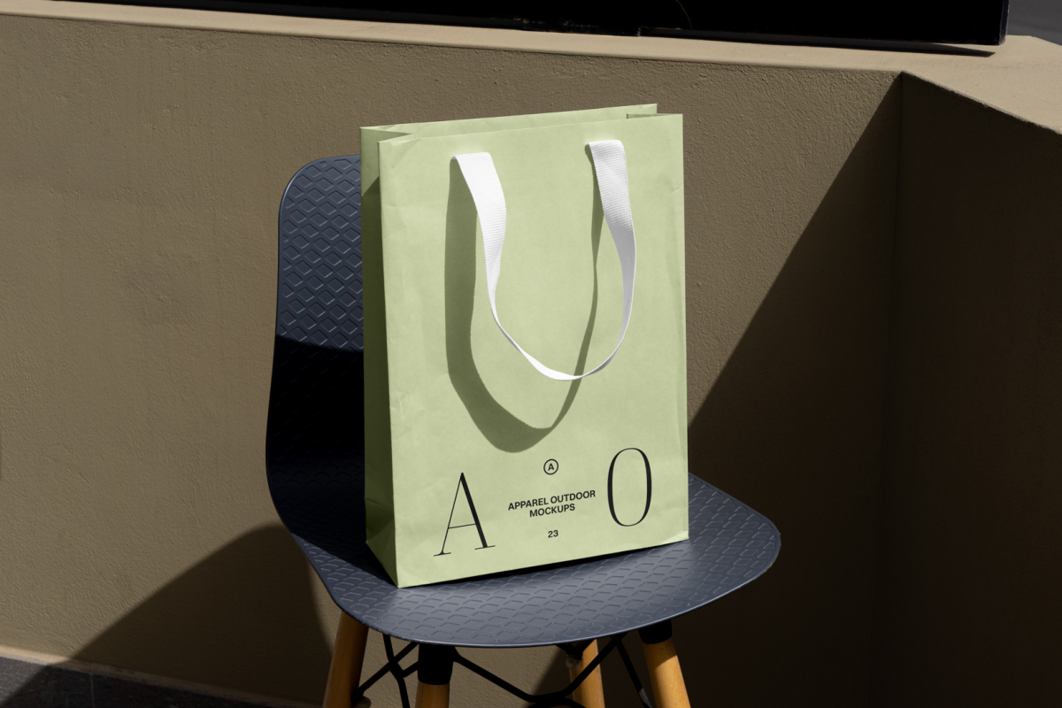 Front view, luxury green Paper Bag Mockup with a white handle and black typographic design on it standing on the navy chair with wooden legs against beige wall.